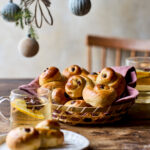 Kurv med nybagte luciabrød serveret på et træbord med varme drikke og julepynt i baggrunden.