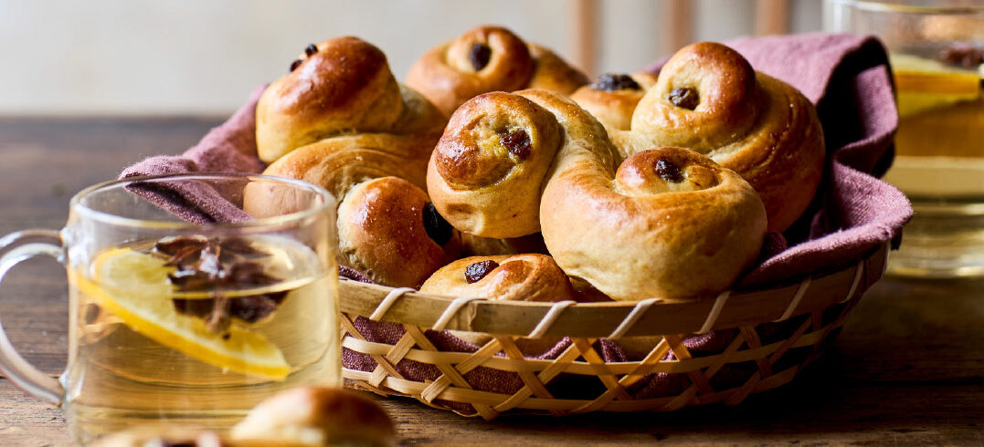 Kurv med nybagte luciabrød serveret på et træbord med varme drikke og julepynt i baggrunden.