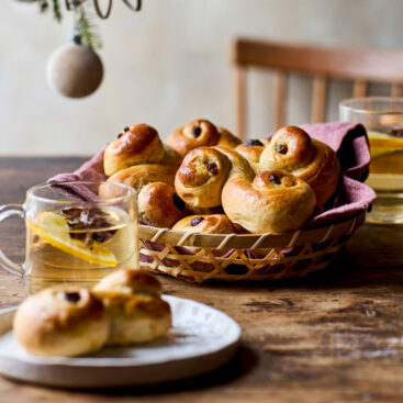 Kurv med nybagte luciabrød serveret på et træbord med varme drikke og julepynt i baggrunden.
