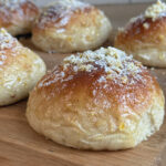 Nybagte juleboller med gylden overflade, penslet med appelsinsirup og drysset med appelsinskal og flormelis, anrettet på et træskærebræt.