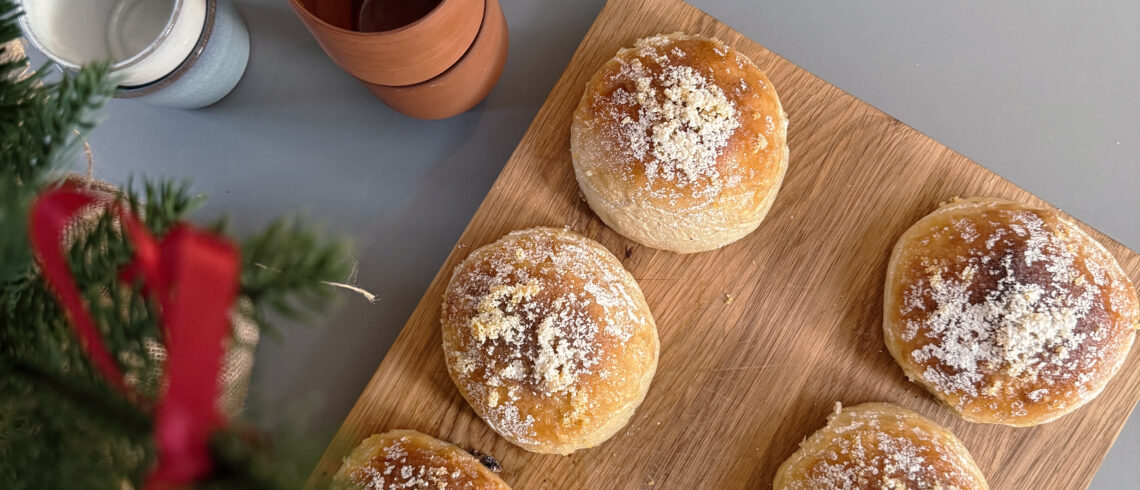 Nybagte juleboller med gylden overflade, penslet med appelsinsirup og drysset med appelsinskal og flormelis, anrettet på et træskærebræt.