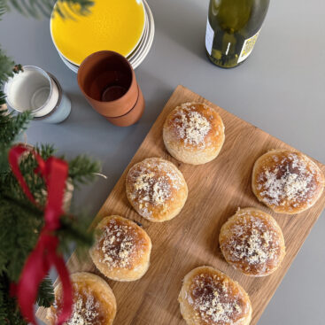 Nybagte juleboller med gylden overflade, penslet med appelsinsirup og drysset med appelsinskal og flormelis, anrettet på et træskærebræt.