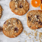 Store julecookies med chokolade, hasselnødder og havreris liggende på en lys marmorbordplade sammen med halve appelsiner og løse havreris.