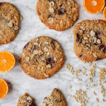 Store julecookies med chokolade, hasselnødder og havreris liggende på en lys marmorbordplade sammen med halve appelsiner og løse havreris.