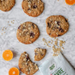 Store julecookies med chokolade, hasselnødder og havreris liggende på en lys marmorbordplade sammen med halve appelsiner og løse havreris.