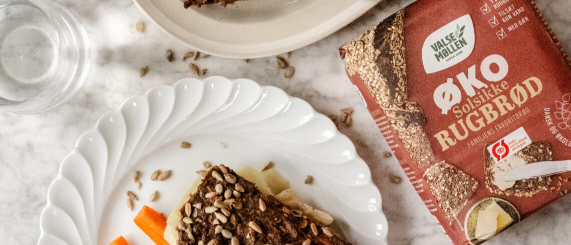 Skolerugbrød med gulerødder og solsikkekerner serveret på tallerken med agurk og gulerodsstave, set oppefra, sammen med pakke Valsemøllen Økologisk Solsikkerugbrød.