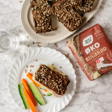 Skolerugbrød med gulerødder og solsikkekerner serveret på tallerken med agurk og gulerodsstave, set oppefra, sammen med pakke Valsemøllen Økologisk Solsikkerugbrød.