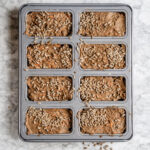 Rugbrødsdej med gulerødder fordelt i portionsforme og drysset med solsikkekerner før bagning.