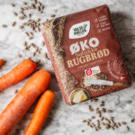 Pakke med Valsemøllen Økologisk Solsikkerugbrød sammen med gulerødder og solsikkekerner på lys marmorbord, set oppefra.